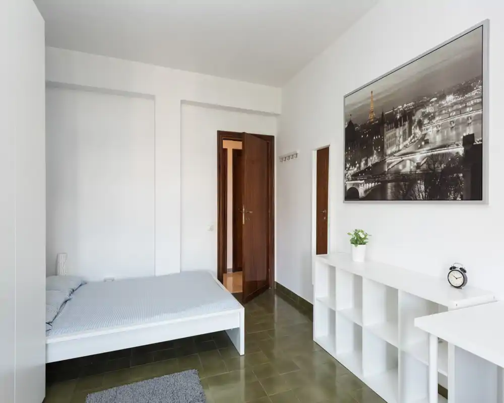 Camera da letto minimalista con letto basso, scaffalatura bianca, biancheria neutra e grande quadro con panorama cittadino; luminosa, pulita e arredata con semplicità.