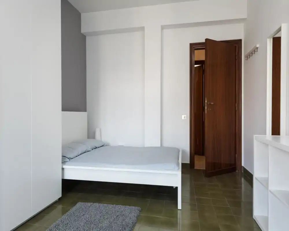 Camera da letto minimalista con letto bianco basso, biancheria grigio chiaro, una lampada da comodino e armadi dalle linee pulite — spazio luminoso, tranquillo e ordinato, perfetto per annunci.