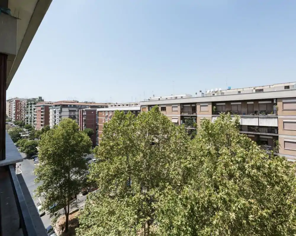 Vista dal balcone soleggiato su alberi rigogliosi e condomini vicini, perfetta per mostrare lo spazio esterno e il panorama urbano.