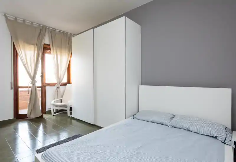 Camera da letto luminosa e minimale con letto matrimoniale, grande armadio bianco e luce naturale dalla porta del balcone — linee pulite e arredamento neutro per un soggiorno tranquillo.