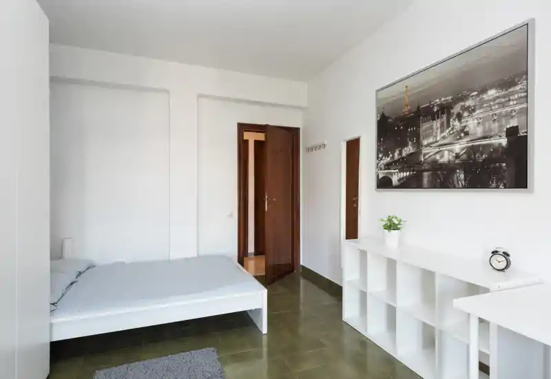 Camera da letto minimalista con letto basso, scaffalatura bianca, biancheria neutra e grande quadro con panorama cittadino; luminosa, pulita e arredata con semplicità.