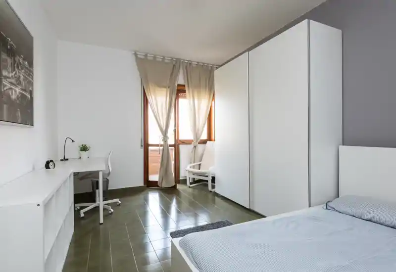 Camera da letto luminosa e minimalista con letto ordinato, grande armadio, scrivania e finestra/porta finestra che procura luce naturale.