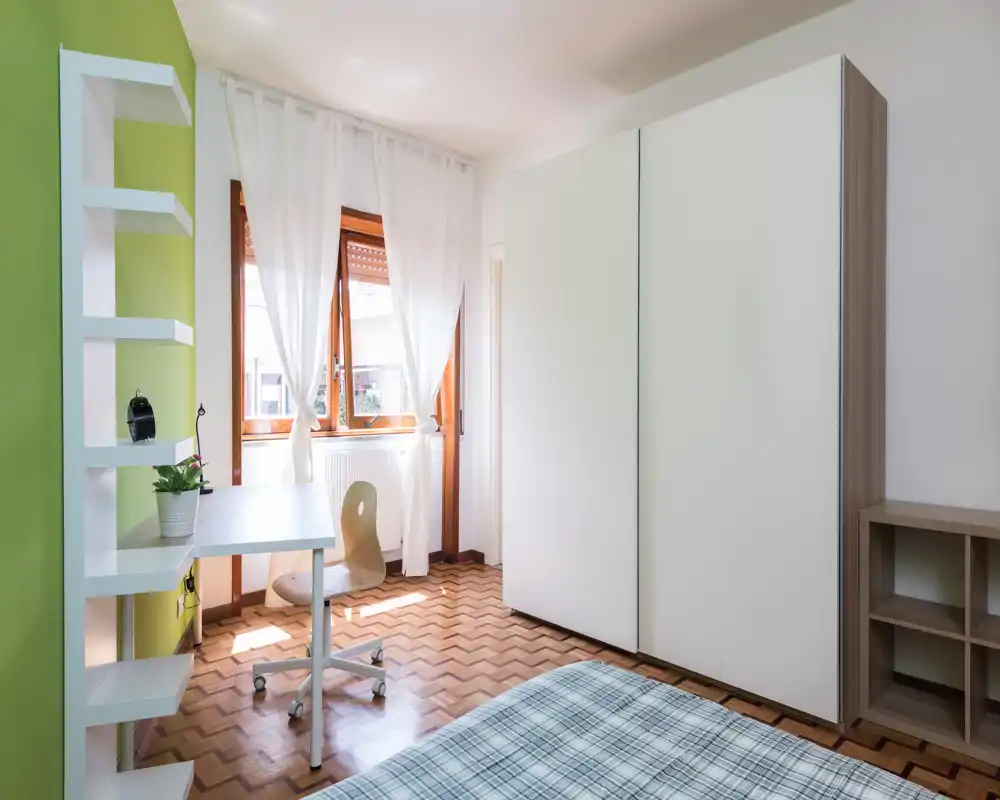 Bright, tidy bedroom with a plaid-covered bed in the foreground, a large white wardrobe, a small desk and chair by the window, and wooden parquet flooring. Natural light and simple modern furnishings create a welcoming, functional space ideal for a single occupant or guest.