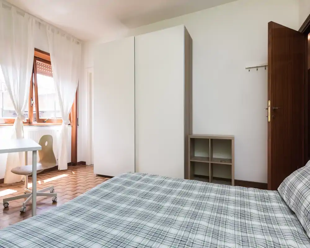 Bright, simple bedroom with a large double bed in foreground, white wardrobe, desk by the window and natural light from sheer curtains.