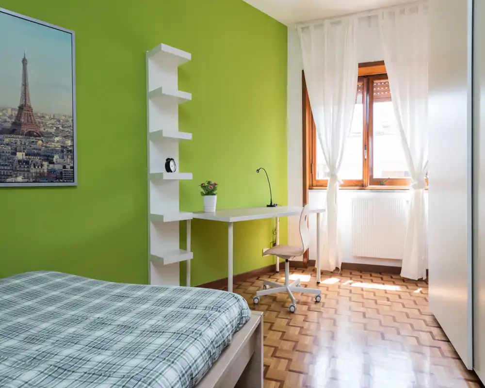 Bright, tidy bedroom with a single bed in the foreground, a study desk by the window and fresh natural light. The green accent wall and parquet floor create a welcoming, modern atmosphere.
