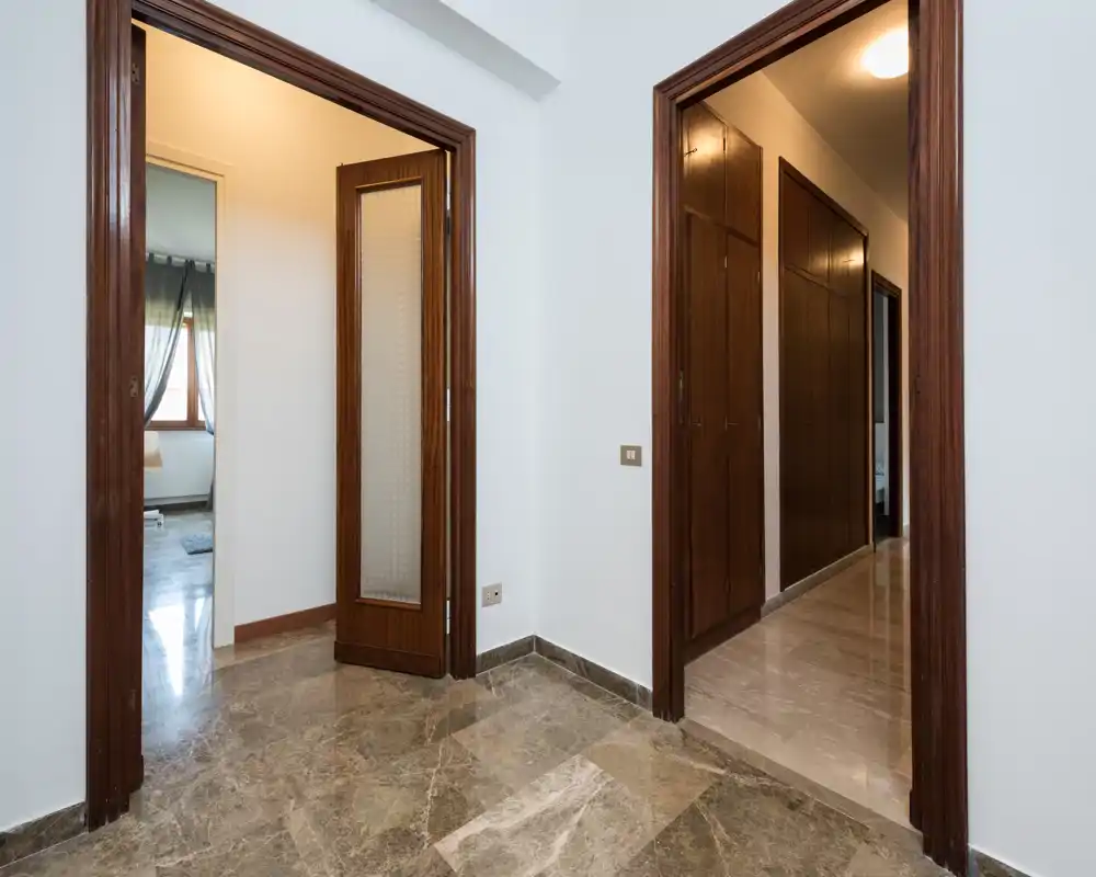 Ingresso/disimpegno luminoso con pavimento in marmo e cornici delle porte in legno che conducono alle stanze adiacenti. Lo spazio è pulito e neutro, ideale per mostrare i collegamenti tra i locali.