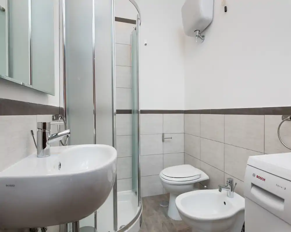 Bagno compatto e moderno con box doccia angolare, lavabo a parete, bidet e wc. Piastrelle neutre e buona illuminazione creano un ambiente pulito e funzionale.