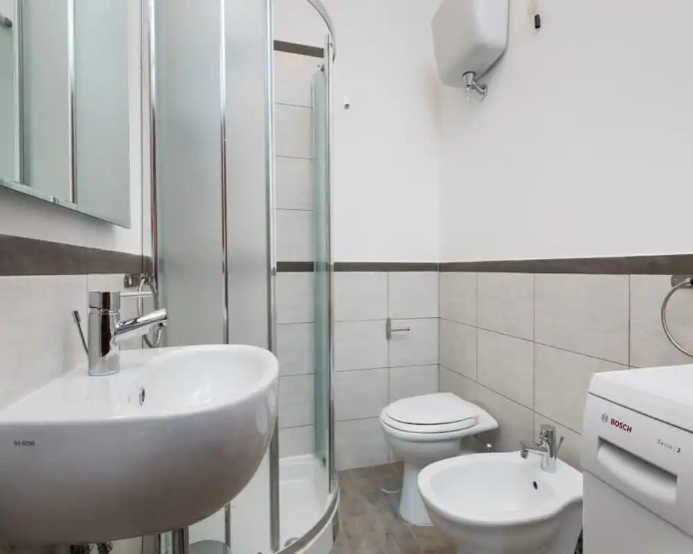 Bagno moderno e pulito con doccia a vetri curva, lavabo, WC e bidet; piastrelle neutre e lavatrice rendono l'ambiente pratico e ben illuminato.