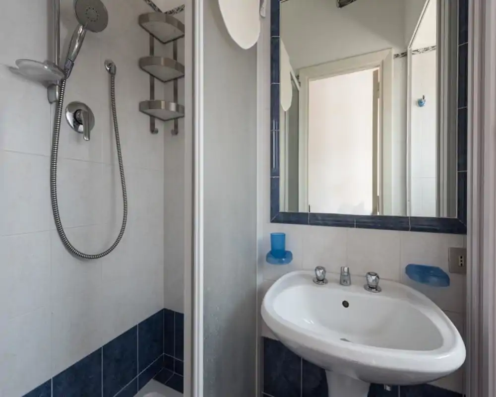 Bagno compatto e luminoso con lavabo a colonna, grande specchio e doccia angolare con doccetta; piastrelle bianche e blu pulite.