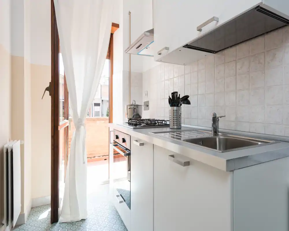 Cucina compatta e luminosa con mobili bianchi, lavello in acciaio e piano cottura a gas che si apre su un piccolo balcone; stile moderno e pulito ideale per appartamenti in città.
