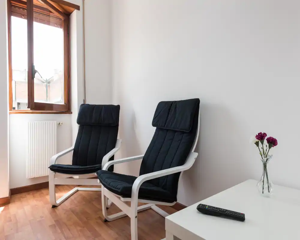Area salotto luminosa e minimal con due poltrone imbottite moderne vicino a una finestra, pavimento in legno e un tavolino con un vaso — ideale per leggere o rilassarsi.