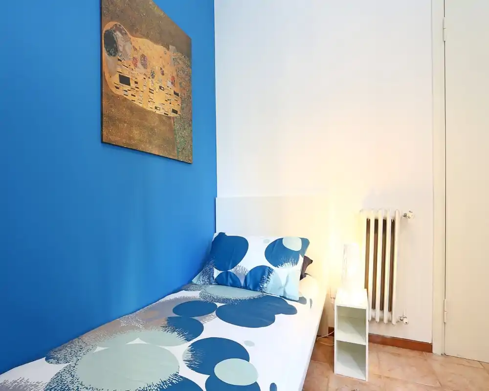 Habitación individual compacta con pared azul llamativa, ropa de cama con estampado y lámpara de mesita — luminosa, ordenada y perfecta para alquileres cortos.
