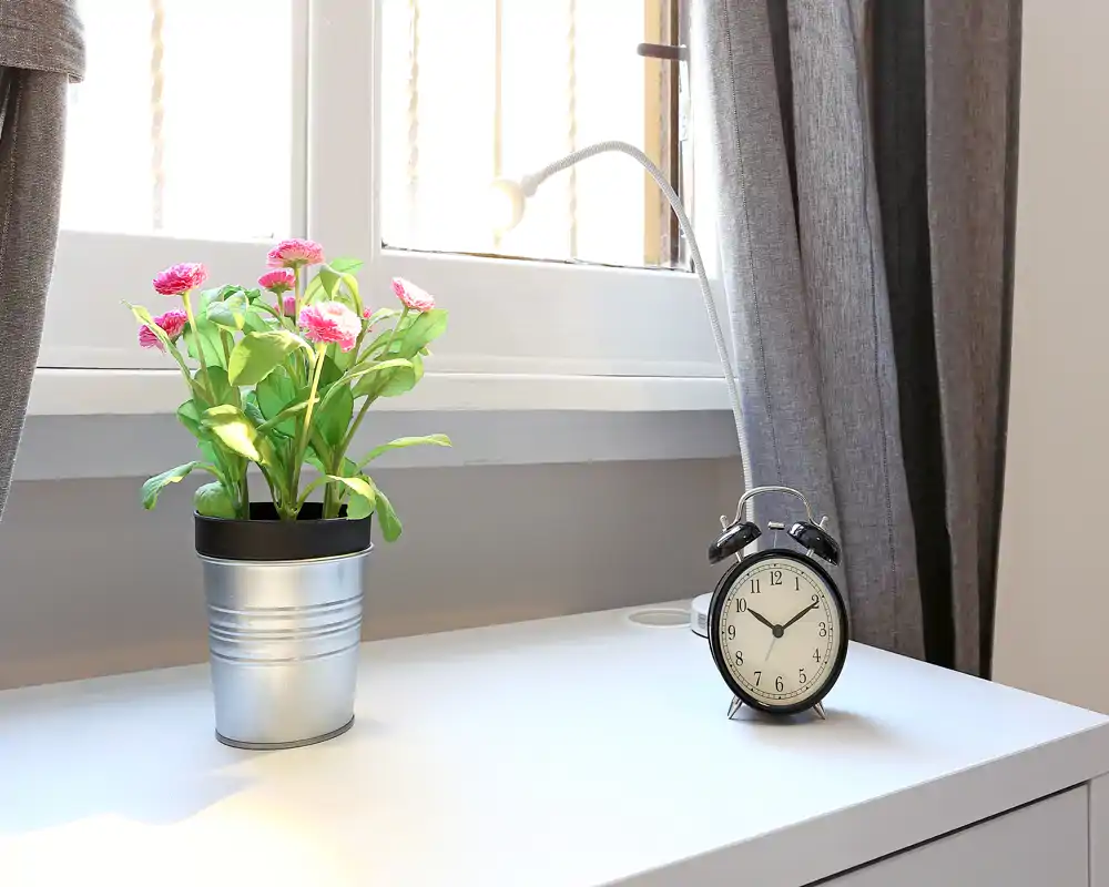 Escena luminosa y minimalista de mesita de noche con planta en maceta, despertador y lámpara de lectura sobre una superficie blanca junto a la ventana — ideal como foto de detalle para destacar la luz y la decoración del dormitorio.