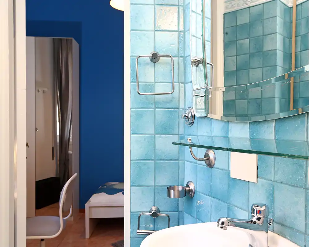 Vista de un baño compacto con paredes de azulejos turquesa, espejo de pared, estante de vidrio y grifería cromada sobre un lavabo blanco de cerámica. La imagen resalta los azulejos modernos y los accesorios con nitidez y buena iluminación.