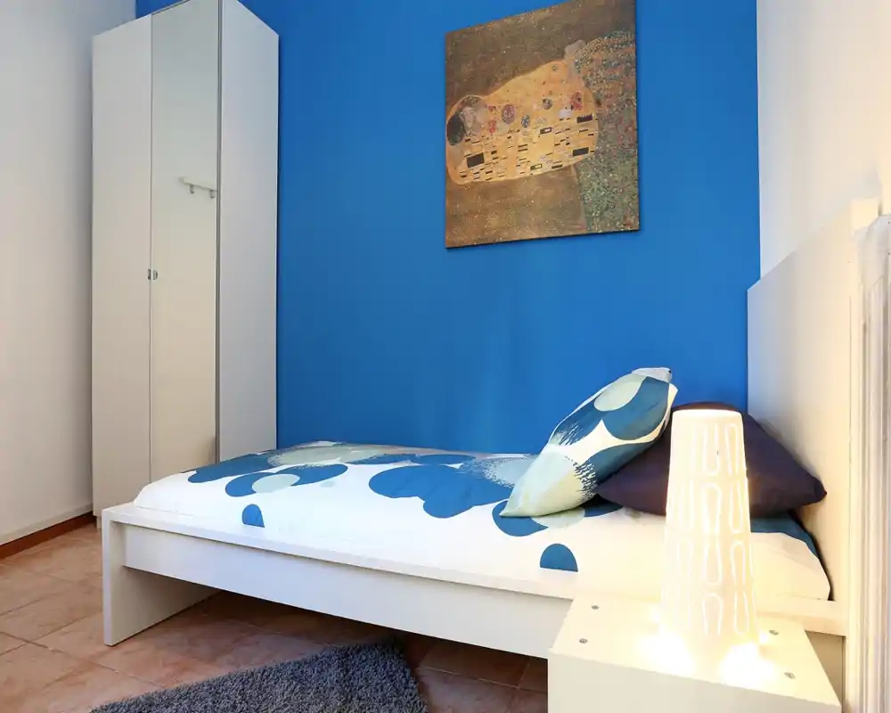 Dormitorio pequeño y moderno con cama individual blanca frente a una pared azul intensa, cojines a juego y lámpara de mesita que aportan calidez.