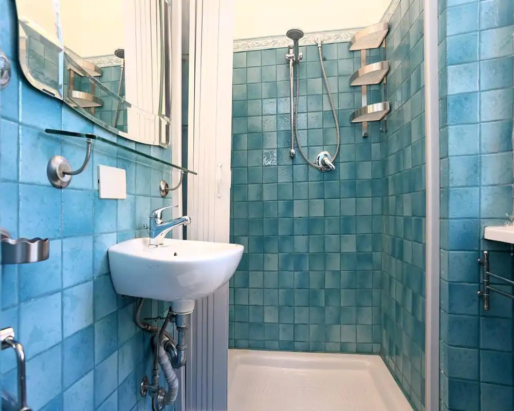 Baño compacto con azulejos turquesa, ducha en esquina con cabezal de mano y pequeño lavabo mural; instalaciones limpias y luminosas, ideal para un alquiler pequeño.