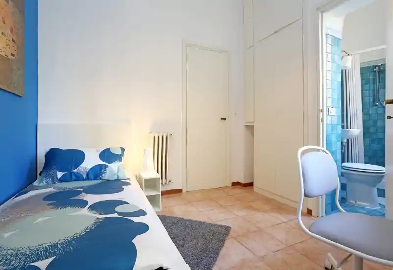 Habitación individual luminosa con ropa de cama azul, lámpara de mesita y alfombra pequeña; baño en suite con azulejos azules visible por la puerta abierta.