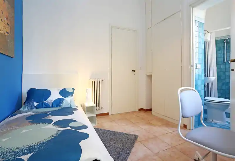 Dormitorio individual luminoso y ordenado con ropa de cama estampada, mesita y lámpara, armario empotrado y baño en suite con azulejos azules.