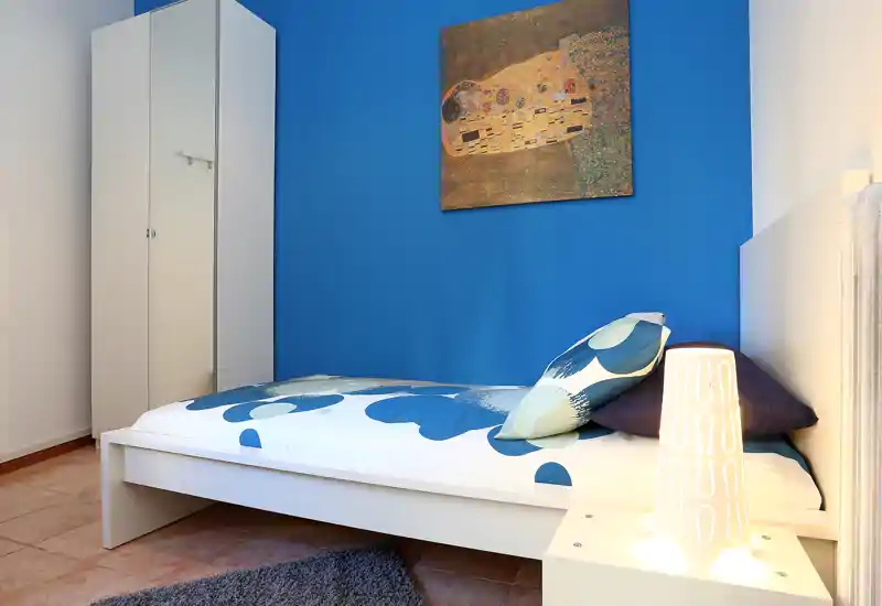 Dormitorio individual moderno y luminoso con cama baja blanca, pared azul de acento y armario minimalista — cojines y lámpara de mesita crean un ambiente acogedor.