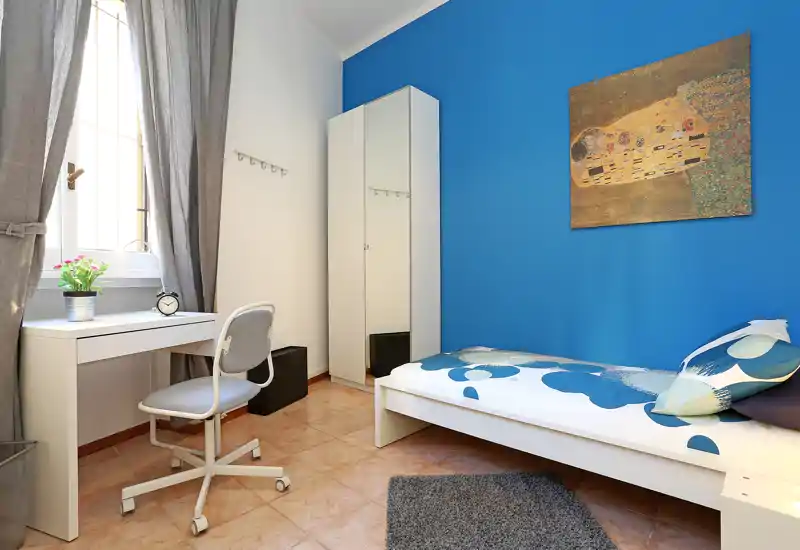 Habitación individual luminosa con cama blanca, cojines estampados y pared azul; zona de estudio junto a la ventana con escritorio y silla, armario con espejo y cuadro decorativo.