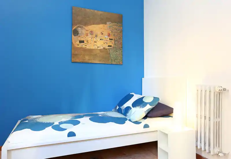 Dormitorio individual compacto y bien presentado con una cama blanca, ropa de cama azul estampada, pared principal azul vivo, cuadro y luz de mesita cálida.