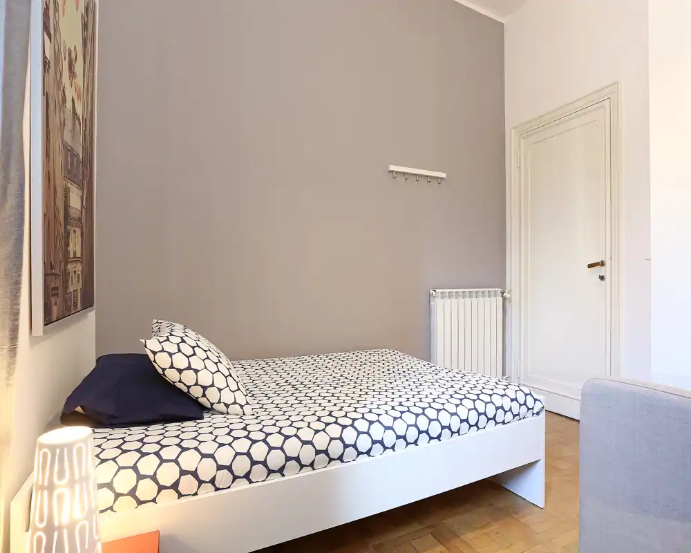 Camera singola moderna e luminosa con copriletto a motivo, lampada da comodino e pareti neutre: ambiente pulito e ben illuminato ideale per brevi soggiorni.