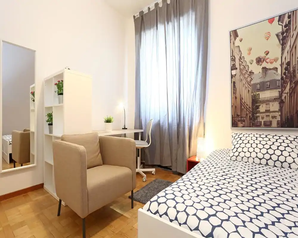 Camera da letto luminosa e curata con letto matrimoniale dal motivo grafico, poltrona comoda, una scrivania vicino alla finestra e quadro decorativo. La luce naturale filtrata dalle tende crea un’atmosfera calda e accogliente.
