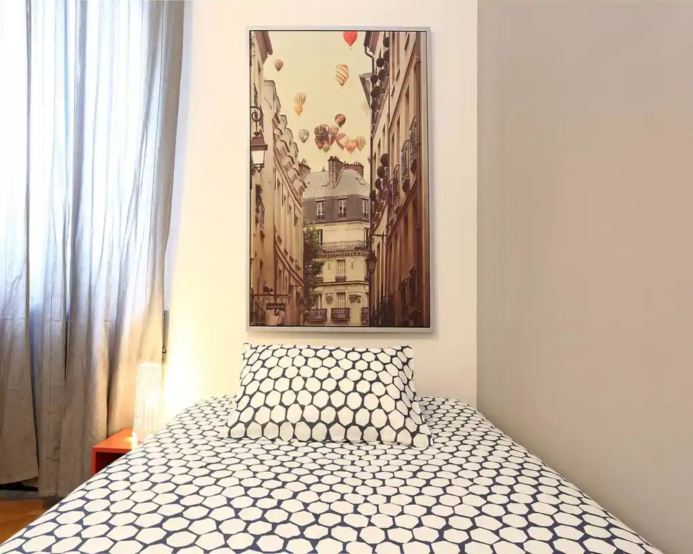 Camera da letto luminosa e accogliente con copriletto a motivo e un cuscino centrale sotto un quadro incorniciato; luce naturale dalla finestra con tenda e una lampada da comodino creano un'atmosfera calda.