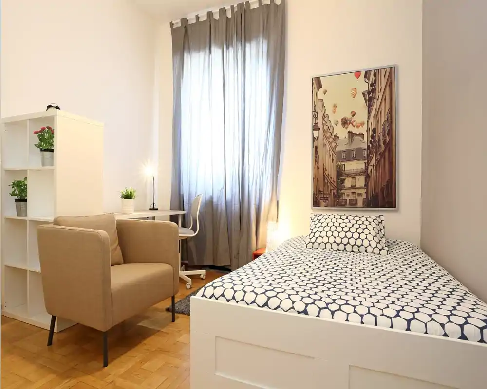 Camera singola luminosa e ben allestita con letto dal motivo grafico, comoda poltrona, scrivania e luce naturale filtrata da una tenda — ideale per un affitto confortevole.