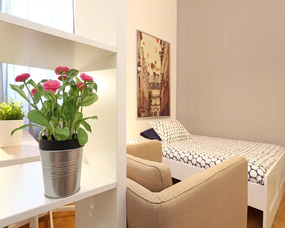 Camera da letto piccola e luminosa con letto singolo con biancheria a motivo, una poltrona confortevole e un quadro decorativo; una libreria con piante aggiunge un tocco caldo e naturale.