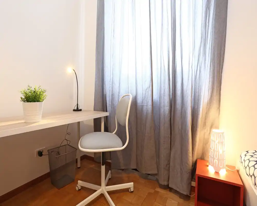 Angolo camera luminoso e minimal con scrivania, sedia e lampada da comodino; si intravede parzialmente il letto sul lato destro dietro tende accoglienti.