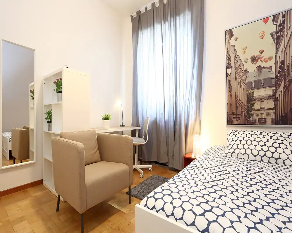Camera da letto luminosa e ben arredata con letto matrimoniale dalla fantasia a motivi, comoda poltrona, scrivania vicino alla finestra e tende morbide che filtrano la luce.