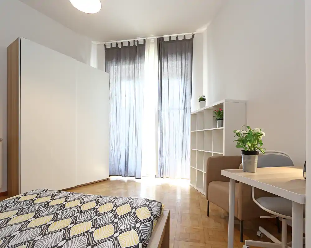Bright, modern bedroom with a patterned double bed in the foreground, large wardrobe, shelving unit and a desk by a sunlit window with sheer curtains.