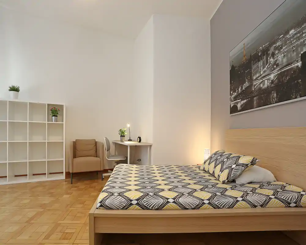 Bright, modern bedroom with a large wooden bed dressed in patterned bedding, a bedside lamp, a small desk and a cozy armchair. Neutral tones and parquet flooring create a calm, welcoming atmosphere ideal for rental listings.