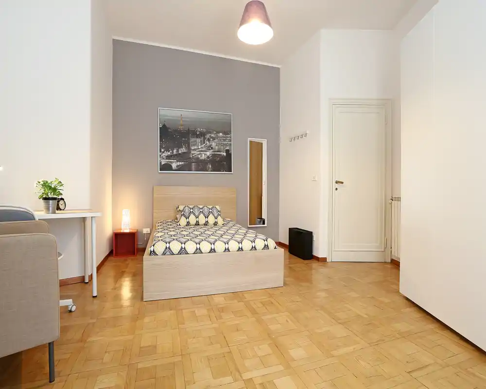 Bright, neatly staged bedroom with a double bed, bedside lamp, desk and wardrobe; warm wooden parquet and tasteful wall art create a welcoming atmosphere.