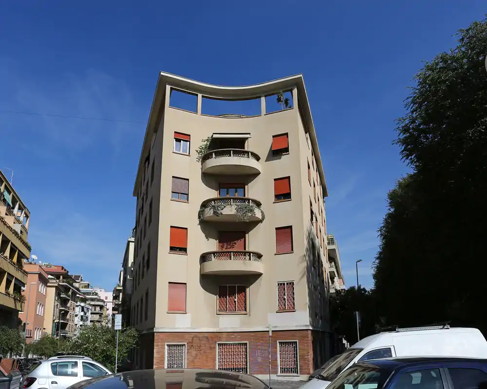 Vista exterior de un edificio de apartamentos con balcones redondeados y persianas rojas, fotografiado desde la calle en un día despejado.