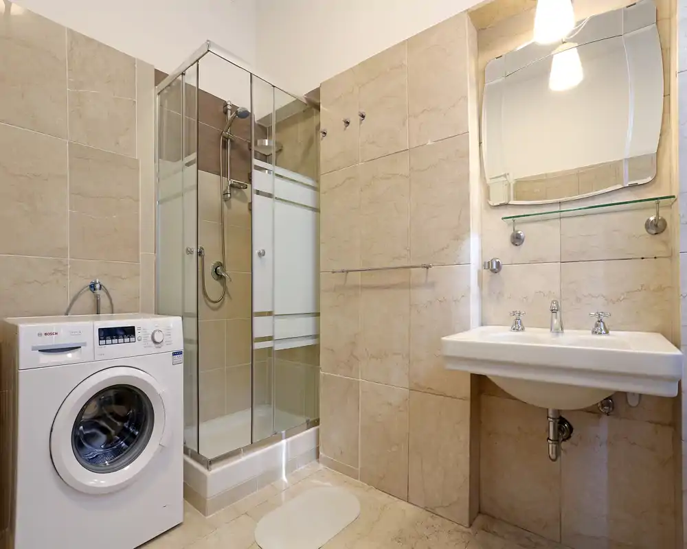 Baño moderno y luminoso con ducha acristalada, lavabo mural, espejo y lavadora. Azulejos beige neutro y accesorios limpios crean un espacio práctico y bien iluminado.