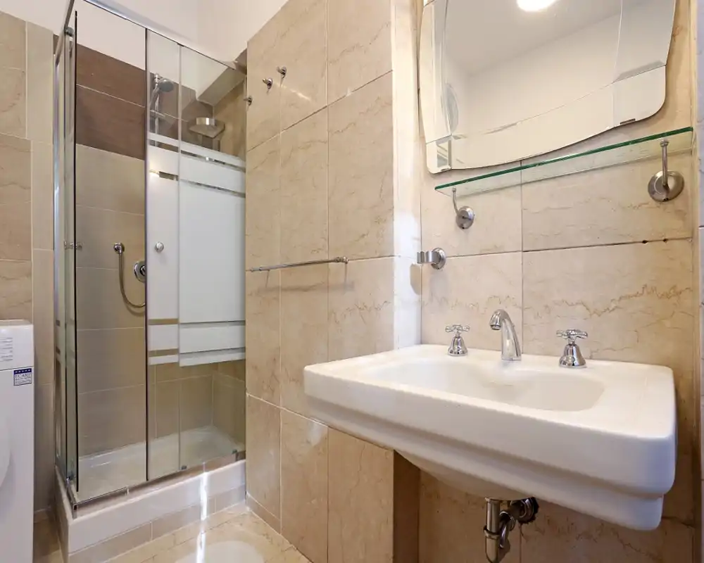 Baño moderno y luminoso con ducha acristalada, lavabo blanco con grifería cromada y azulejos beige efecto mármol.