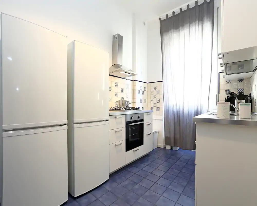 Cocina moderna y luminosa con suelo de baldosas azules, armarios blancos, dos frigoríficos y horno empotrado; salpicadero con motivos y una gran ventana con cortina aportan carácter y buena luz natural.