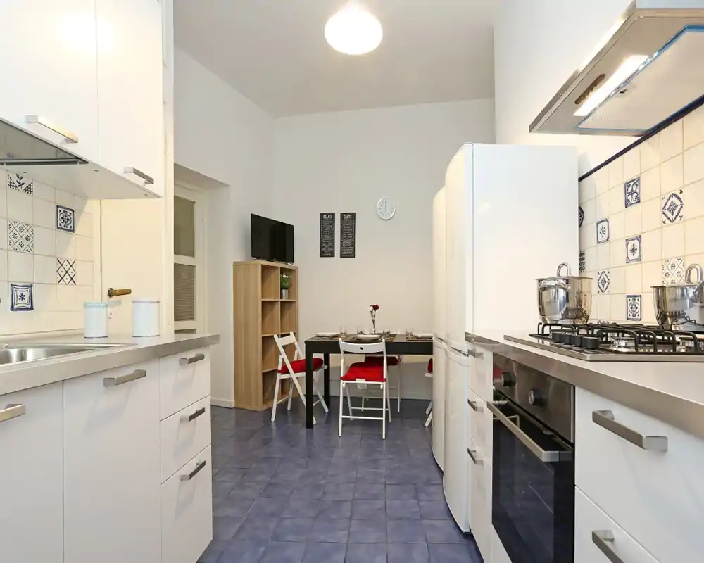 Cocina moderna y luminosa con muebles blancos, placa de gas y horno, salpicadero de azulejos decorativos y una pequeña mesa de comedor — ordenada y bien iluminada.