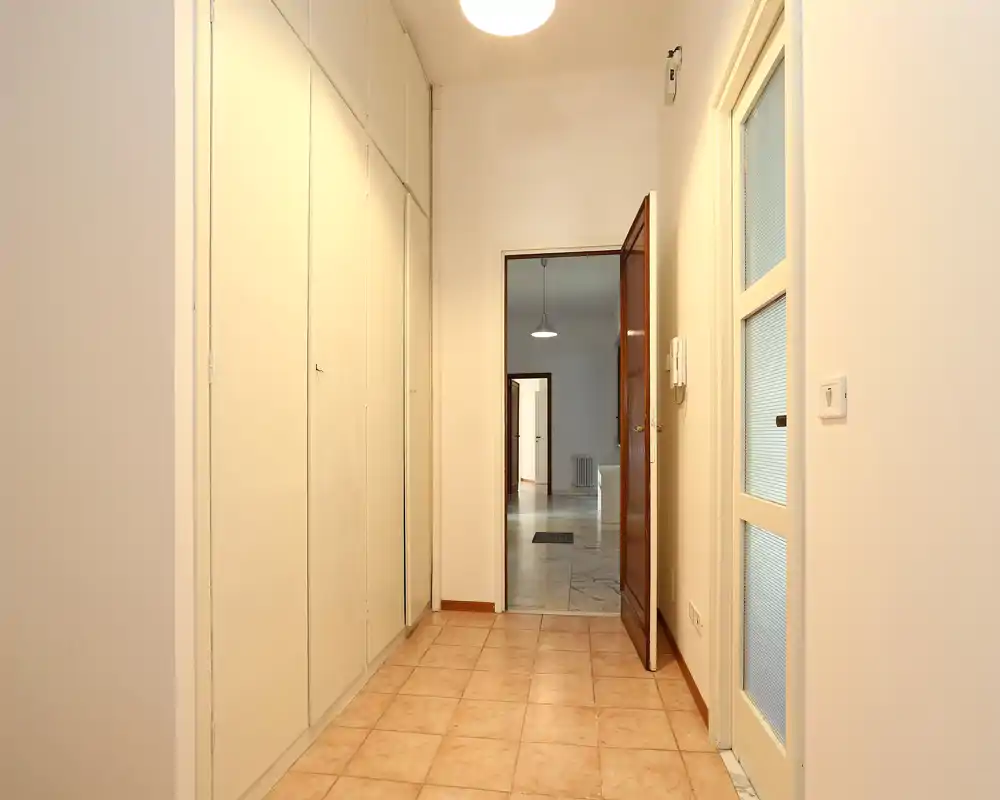 Recibidor luminoso y ordenado con armarios empotrados a la izquierda y una puerta que da al salón. Iluminación cálida en el techo y suelo de baldosas que aportan una impresión acogedora.