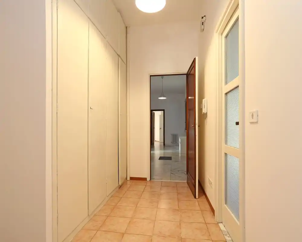 Vestíbulo luminoso con suelo de baldosas, armarios empotrados a la izquierda, puerta de madera abierta al salón y puerta interior acristalada a la derecha.