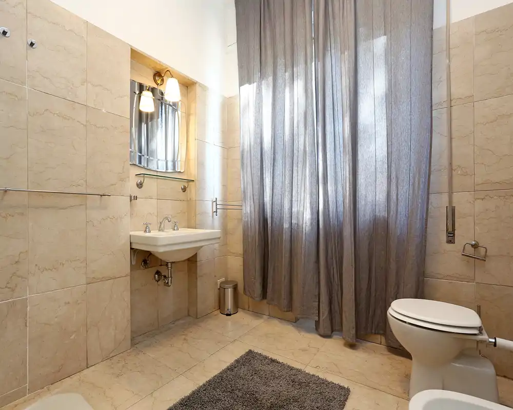 Baño luminoso con azulejos, lavabo de pared, espejo con luces, inodoro y cortina a toda altura para mayor privacidad.