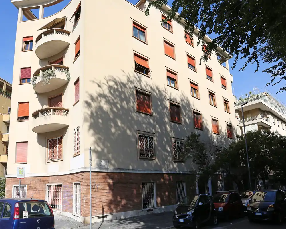 Vista exterior de un edificio residencial de varios pisos con balcones redondeados, ventanas con persianas y coches aparcados en la calle, capturada a plena luz del día.