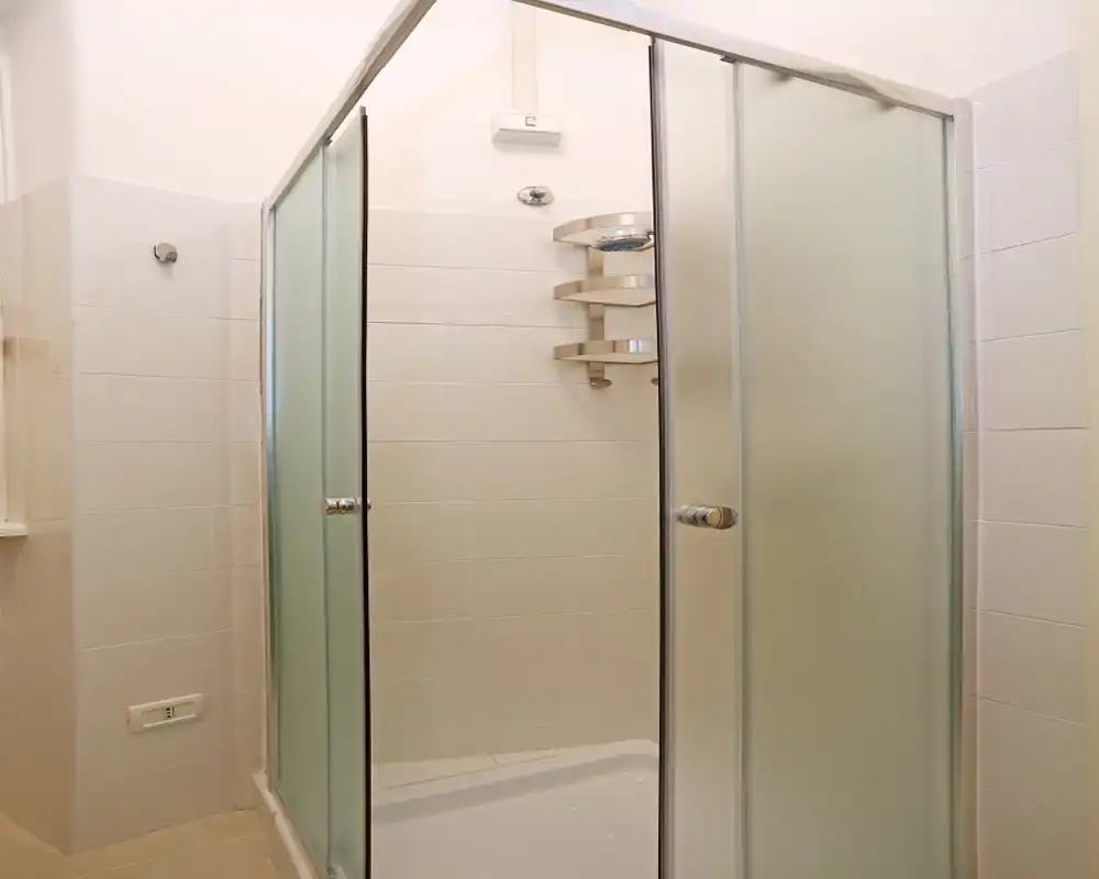 Ducha compacta y moderna con puertas correderas de vidrio esmerilado, paredes con azulejos blancos y estantería de esquina cromada.