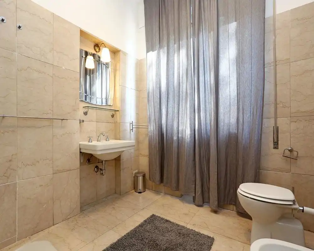 Baño alicatado y limpio con lavabo suspendido, espejo con apliques, inodoro y una gran ventana con cortina que aporta luz natural suave.