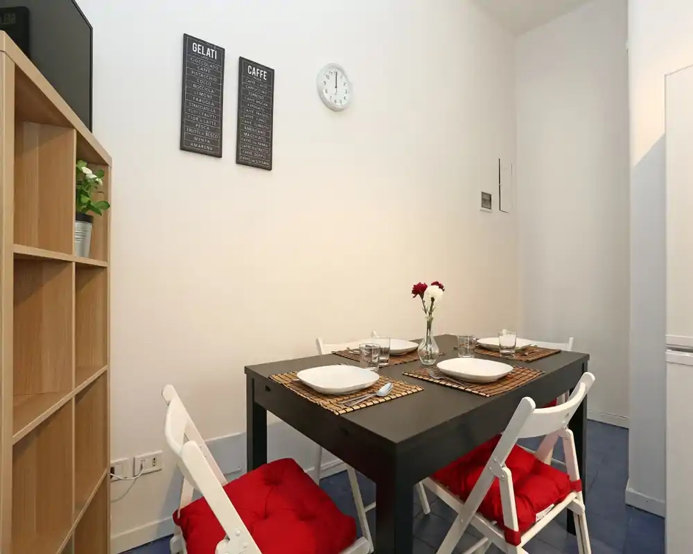 Zona de comedor con estilo: mesa rectangular oscura para cuatro, sillas plegables blancas con cojines rojos y un sencillo centro floral — luminosa, ordenada y acogedora.