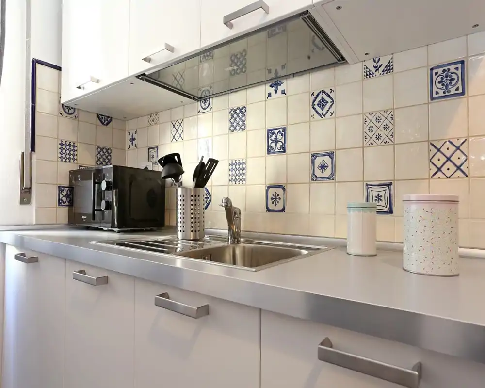 Cocina compacta y luminosa con fregadero de acero inoxidable, salpicadero de azulejos decorativos y electrodomésticos sobre la encimera — un área de preparación ordenada y funcional.