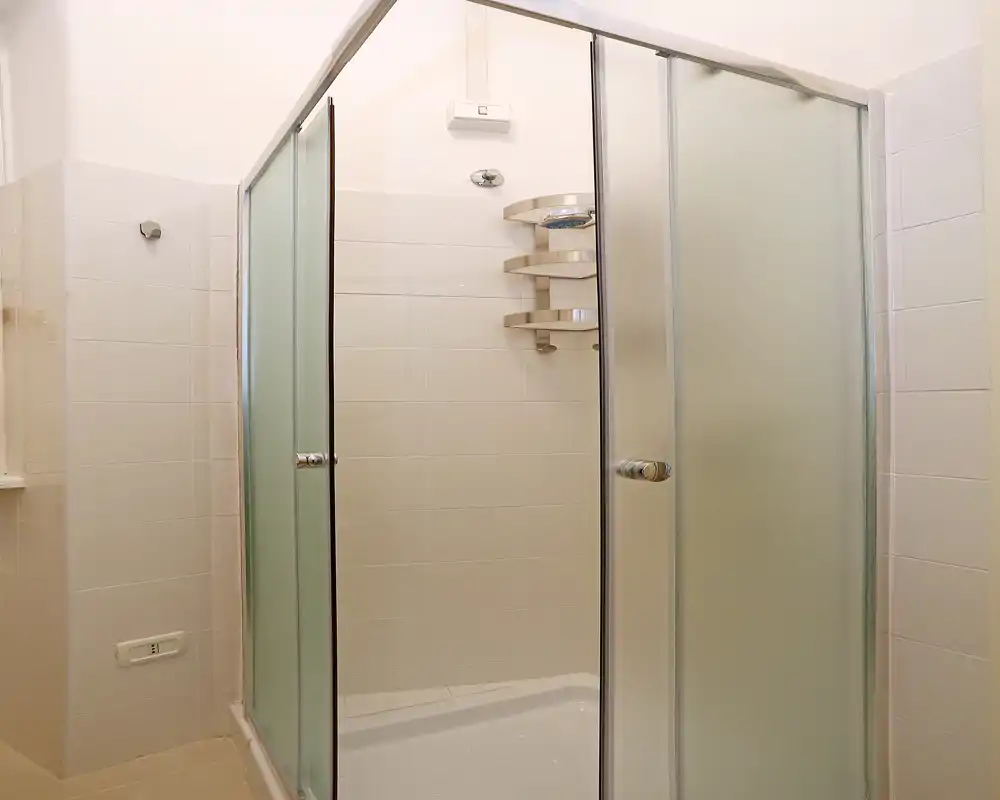 Ducha moderna y luminosa con puertas correderas de vidrio esmerilado, paredes alicatadas y estantería de esquina para artículos de baño.