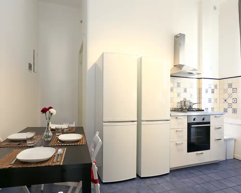 Cocina luminosa y totalmente equipada con una mesa de comedor compacta. Muebles blancos, dos frigoríficos, horno y un salpicadero con dibujos crean un espacio práctico y acogedor, ideal para estancias cortas.